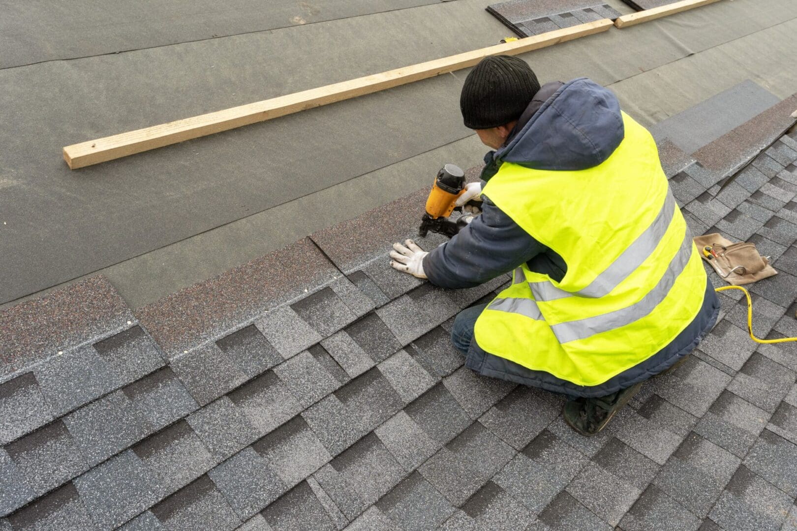 Worker installing asphalt shingles on a roof, wearing a yellow safety vest.